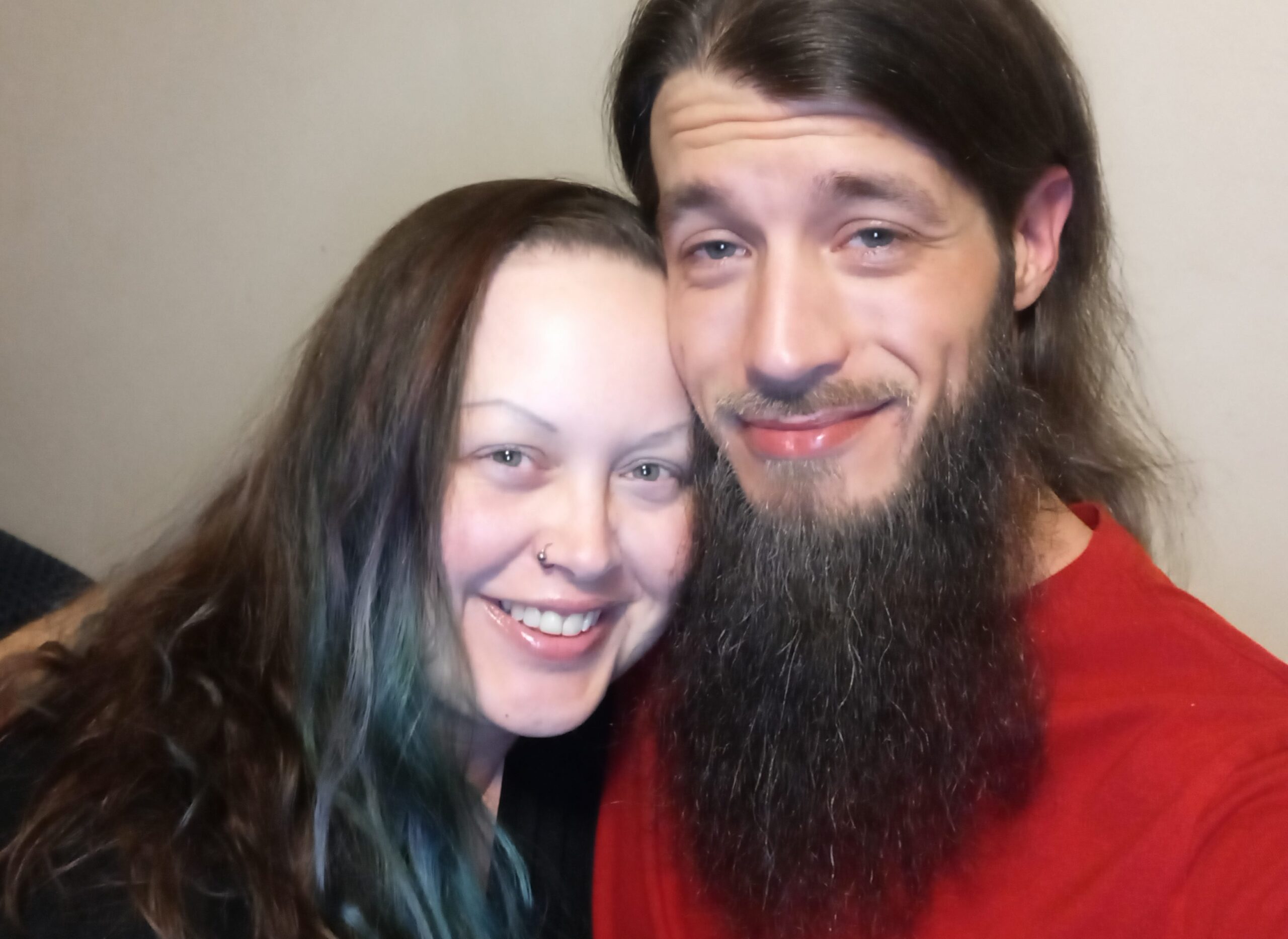 Selfie of David and Didi. David is wearing a bright red shirt and has long, dark brown hair and a long, dark brown beard with silver streaks. Didi is wearing a black shirt and has long, slightly wavy hair that is lighter brown with green and purple streaks. David has his arm around Didi and Didi is leaning his head on David's shoulder. The sides of their faces are touching, and they have big, relaxed smiles on their faces.