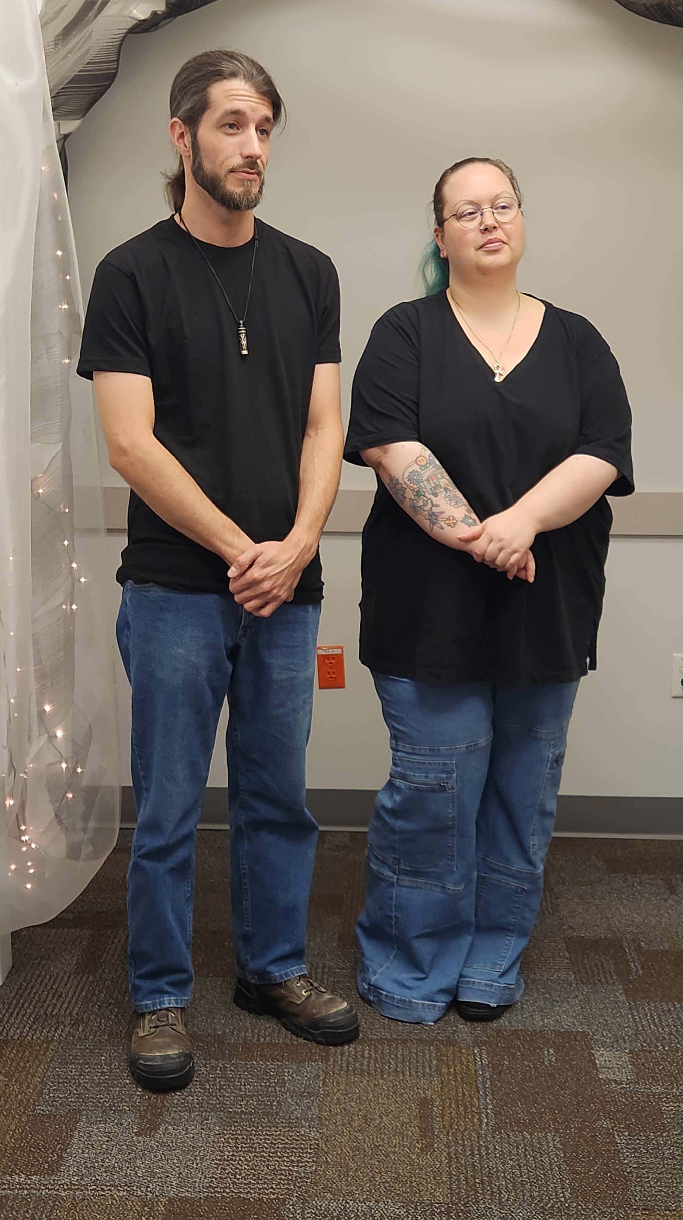 Picture of David and Didi at their wedding. They are both wearing blue jeans, black T-shirts, and boots, their long hair tied back in ponytails. They are standing next to each other in almost the exact same pose. Straight and proud, arms in front of their bodies, left arms holding right wrists. Heads slightly tilted, looking next to the camera.