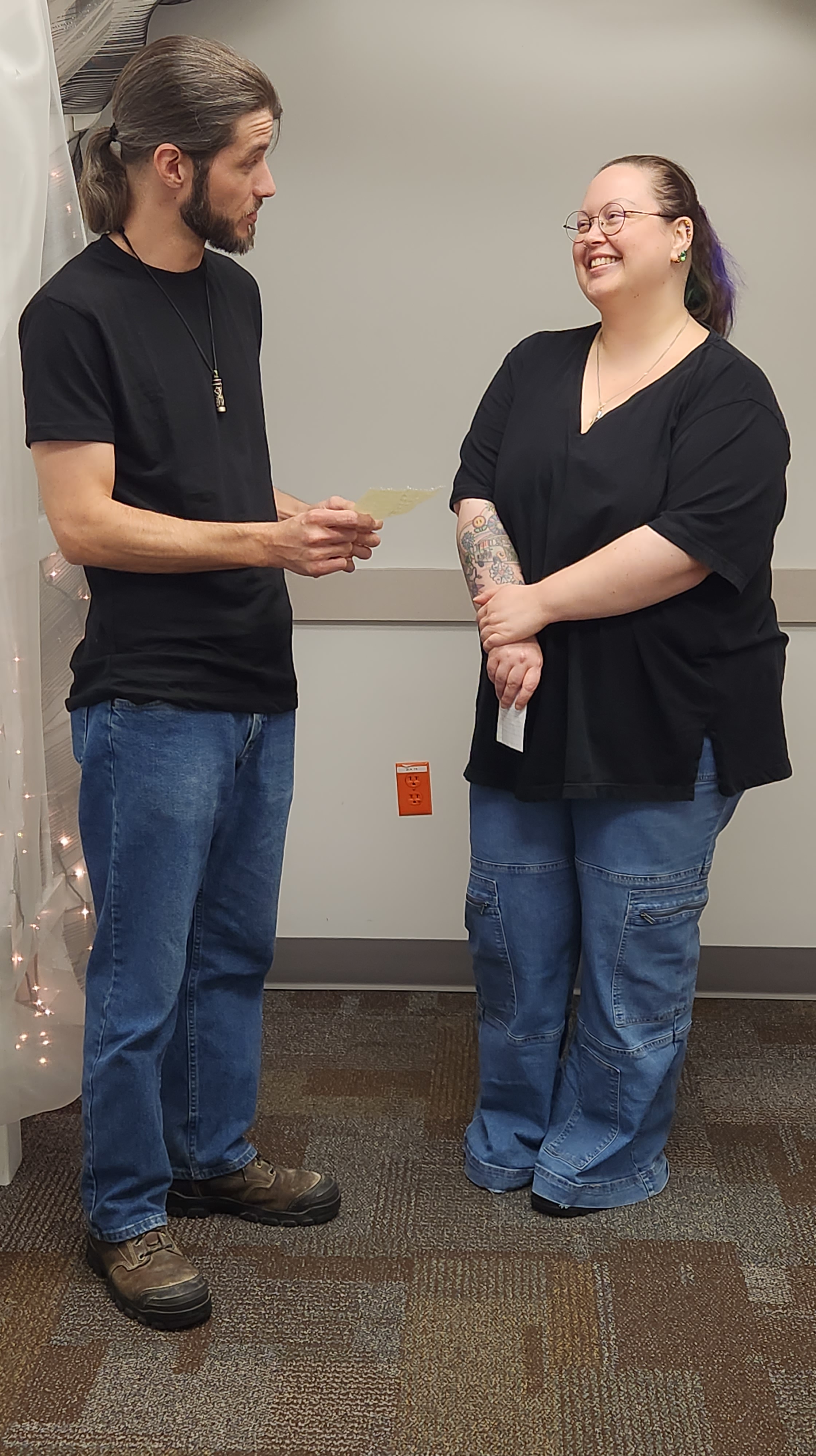 Picture of David and Didi at their wedding. They are both wearing blue jeans, black T-shirts, and boots, their long hair tied back in ponytails. They are facing each other, both holding a small piece of paper. David is reading from his. Didi has a huge smile on his face.