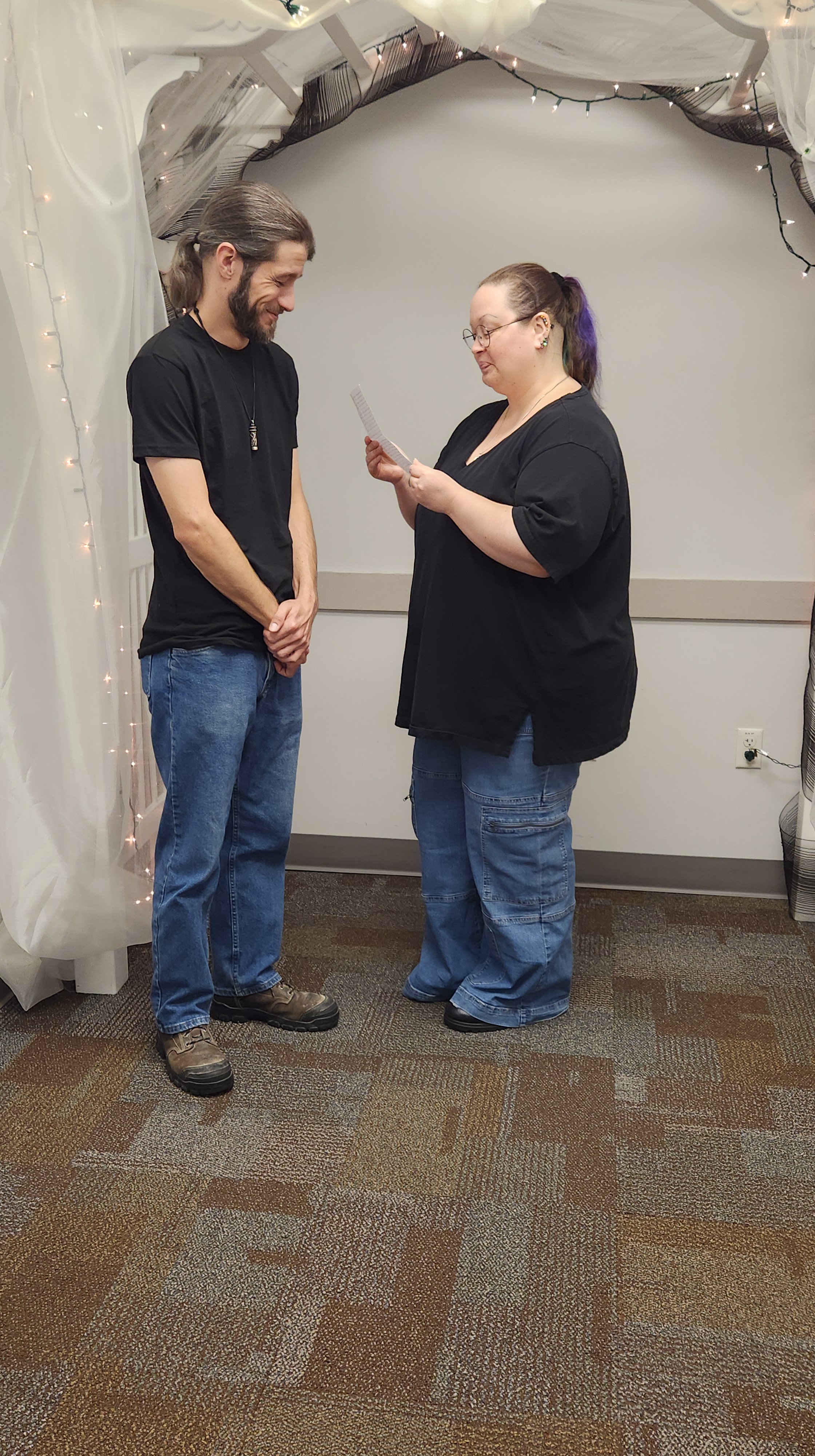 Picture of David and Didi at their wedding. They are both wearing blue jeans, black T-shirts, and boots, their long hair tied back in ponytails. They are facing each other. Didi is reading from a small piece of paper. David is visibly giggling.