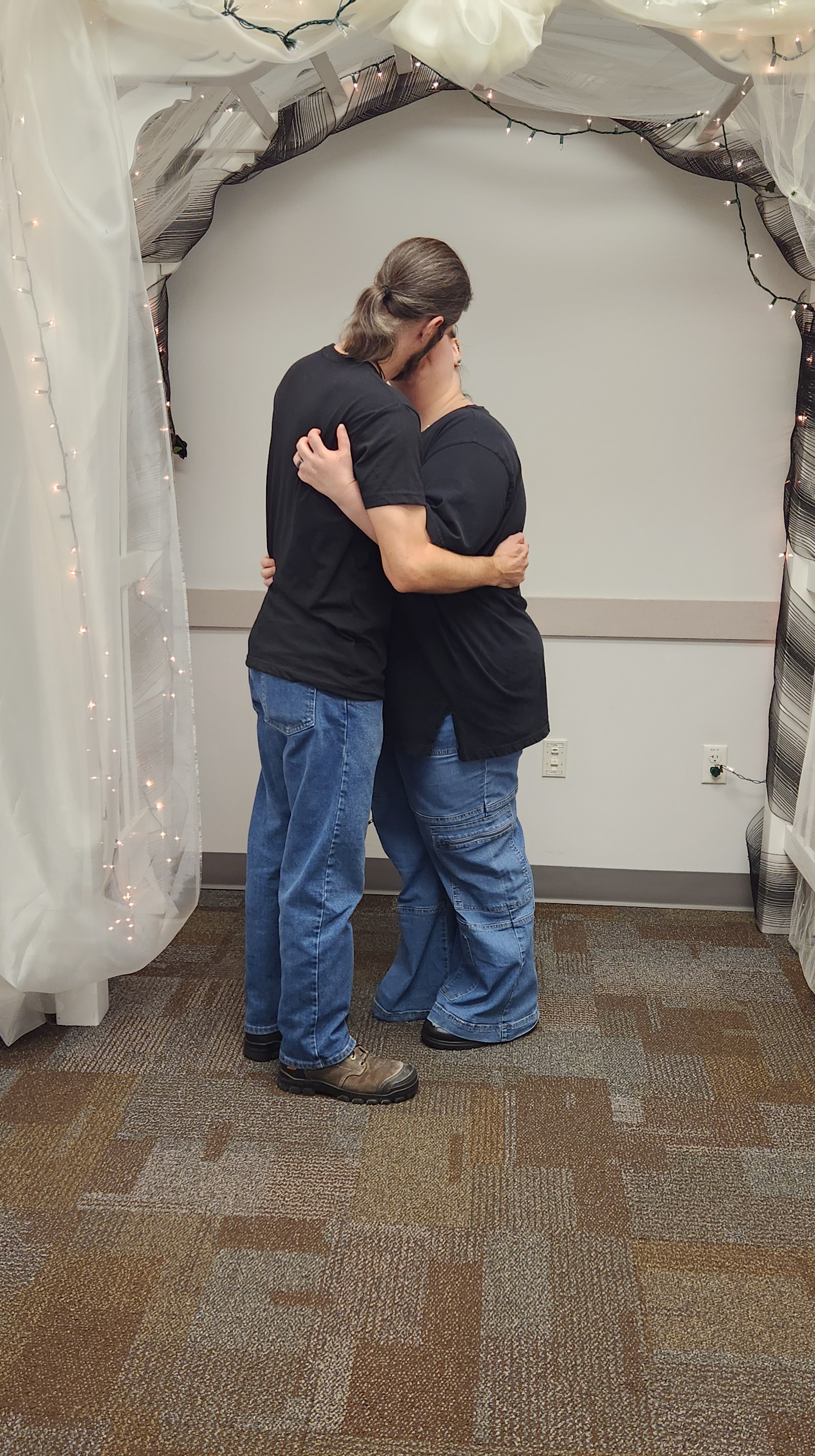 Picture of David and Didi at their wedding. They are both wearing blue jeans, black T-shirts, and boots, their long hair tied back in ponytails. They are holding each other tightly, kissing because they just got married!