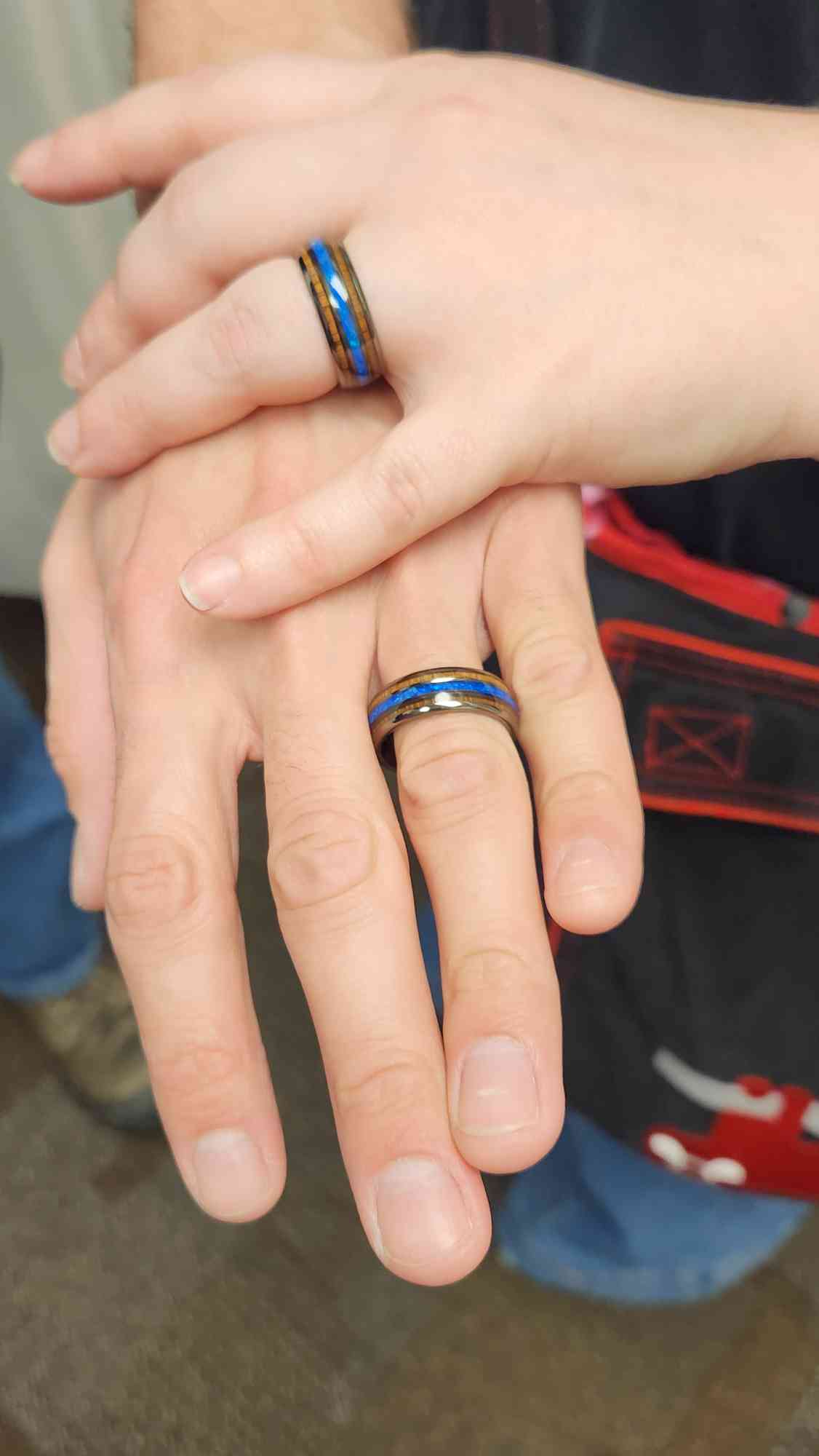 Picture of Didi's small hand on top of David's big hand, showing off their wedding rings. The rings are tungsten with alternating inlaid bands of wood, blue opal, and wood again.