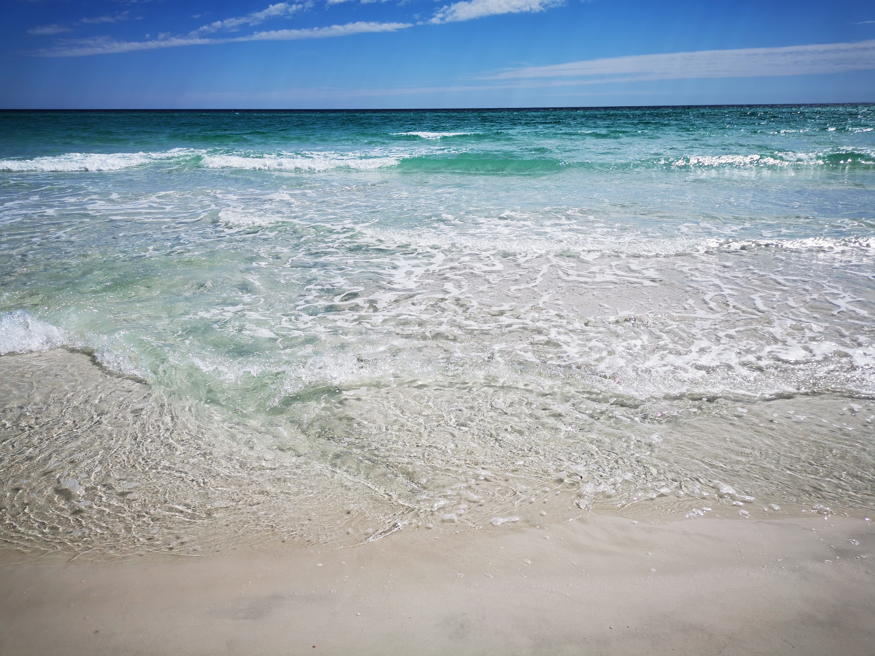Close up picture of the Atlantic ocean, as seen from the very edge of Pensacola Beach. The water nearly touches the camera, and has various colors of green, blue, turquoise.