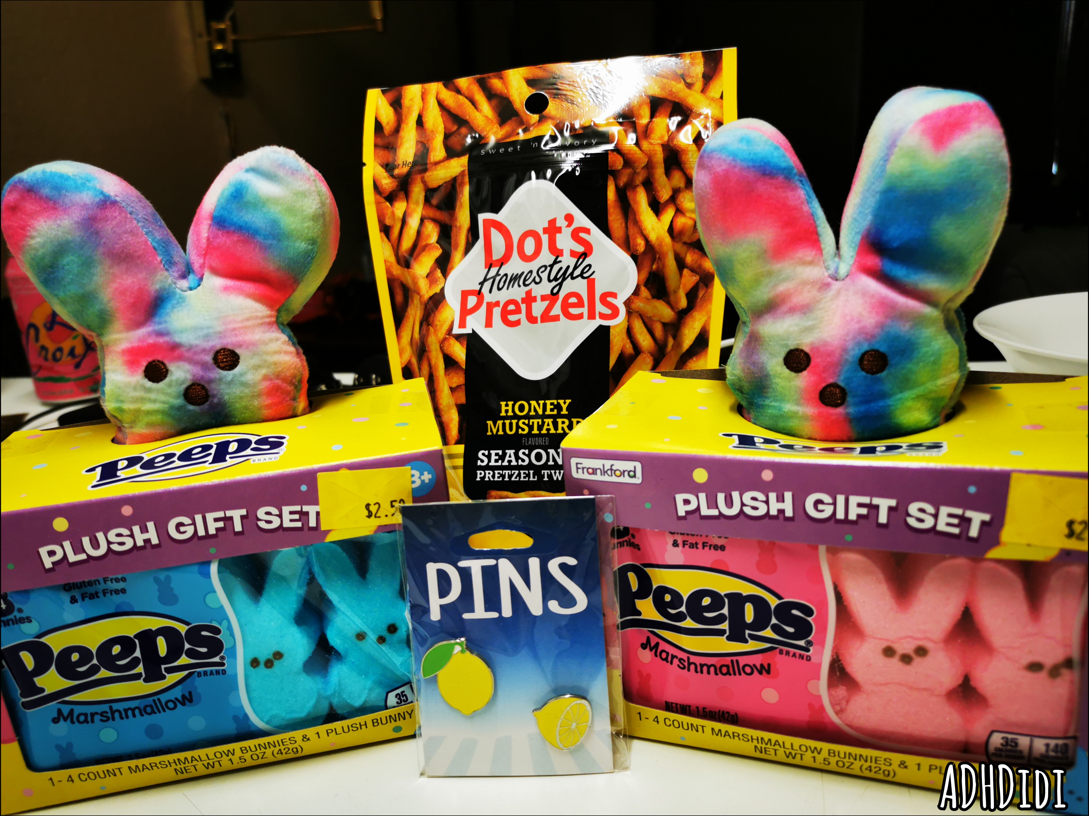 A picture of a collection of gifts and snacks. A small bag of Dot's honey mustard pretzels, two pins depicting a whole lemon and half a lemon, and two gift sets from Peeps. Both gift sets have four edible bunny shaped Peeps, pink in one box and blue in the other. They also contain a tiny rainbow colored Peeps bunny plushy.
