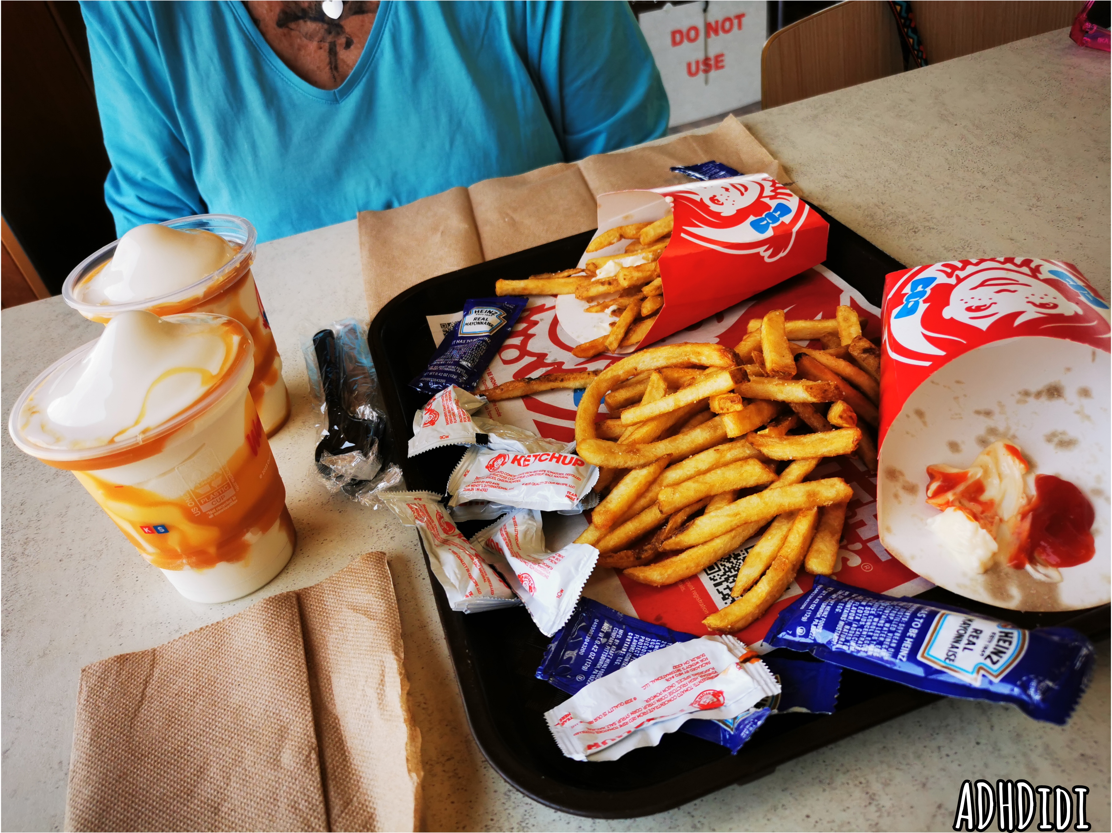 Picture of a slightly chaotic food situation at Wendy's. Fries and way too many ketchup and mayo packets scattered around a black tray. Two caramel frosties standing next to it, already melting. A small part of Mem in a light blue shirt is visible across the table.